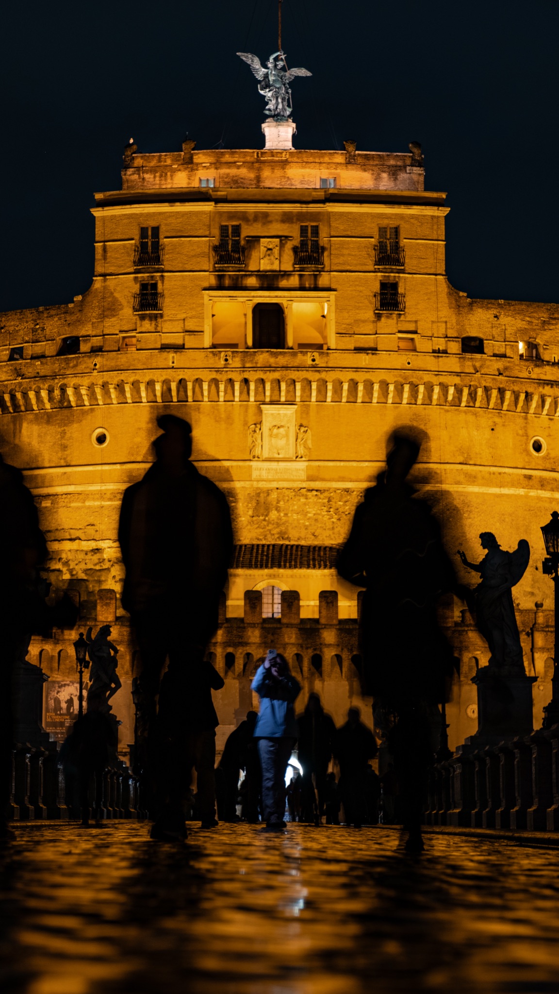 Blurred silhouettes crossing the bridge before floodlit Castel Sant'Angelo, Rome, at night — photograph by Andrea Rubbi