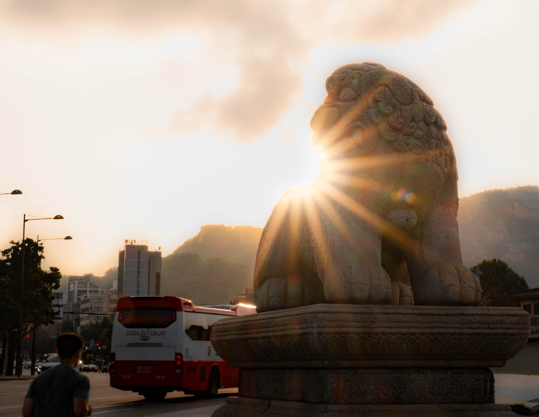 Stone guardian lion backlit by a golden-hour sunstar beside a city bus in Seoul — photograph by Andrea Rubbi